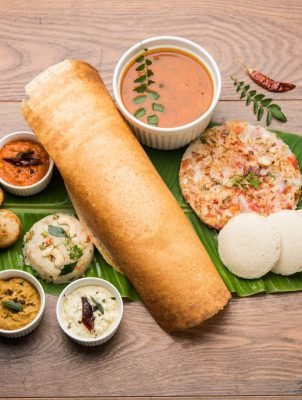 Group of South Indian food like Masala Dosa, Uttapam, Idli or idly, Wada or vada, sambar, appam, semolina halwa, upma served over banana leaf with colourful chutneys, selective focus